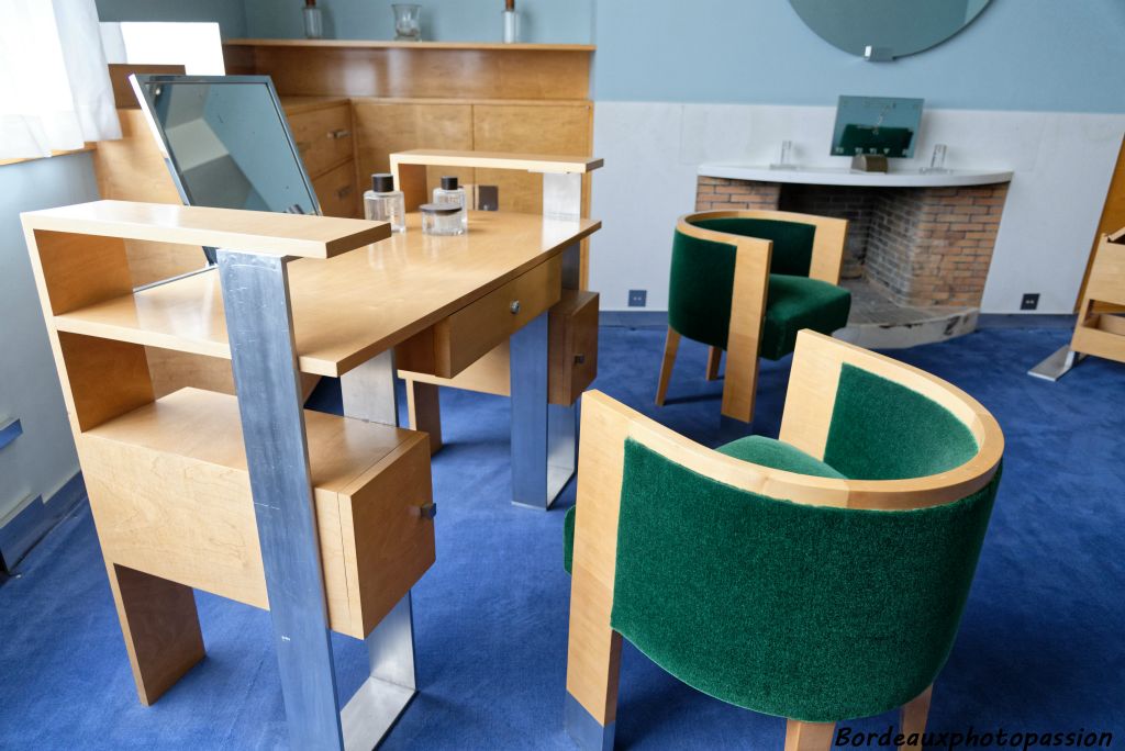 Le mobilier est moderne, très géométrique comme le reste de la villa. On peut noter la polychromie de la pièce : velours vert des sièges, bleu ciel des murs, blondeur des meubles et moquette bleu marine.